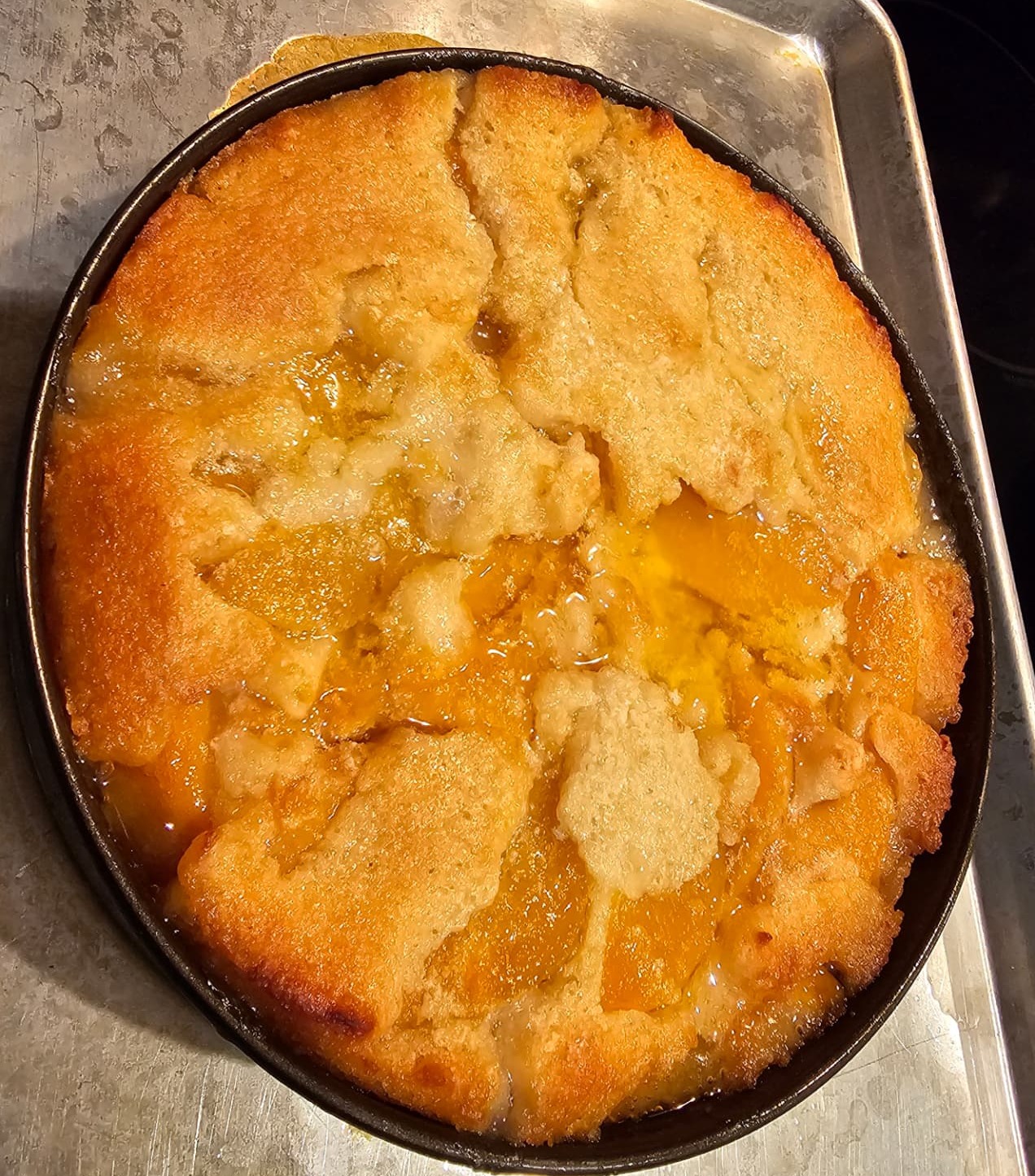 A golden-brown peach cobbler baked in a round skillet, with peach slices and glossy syrup bubbling through the crust