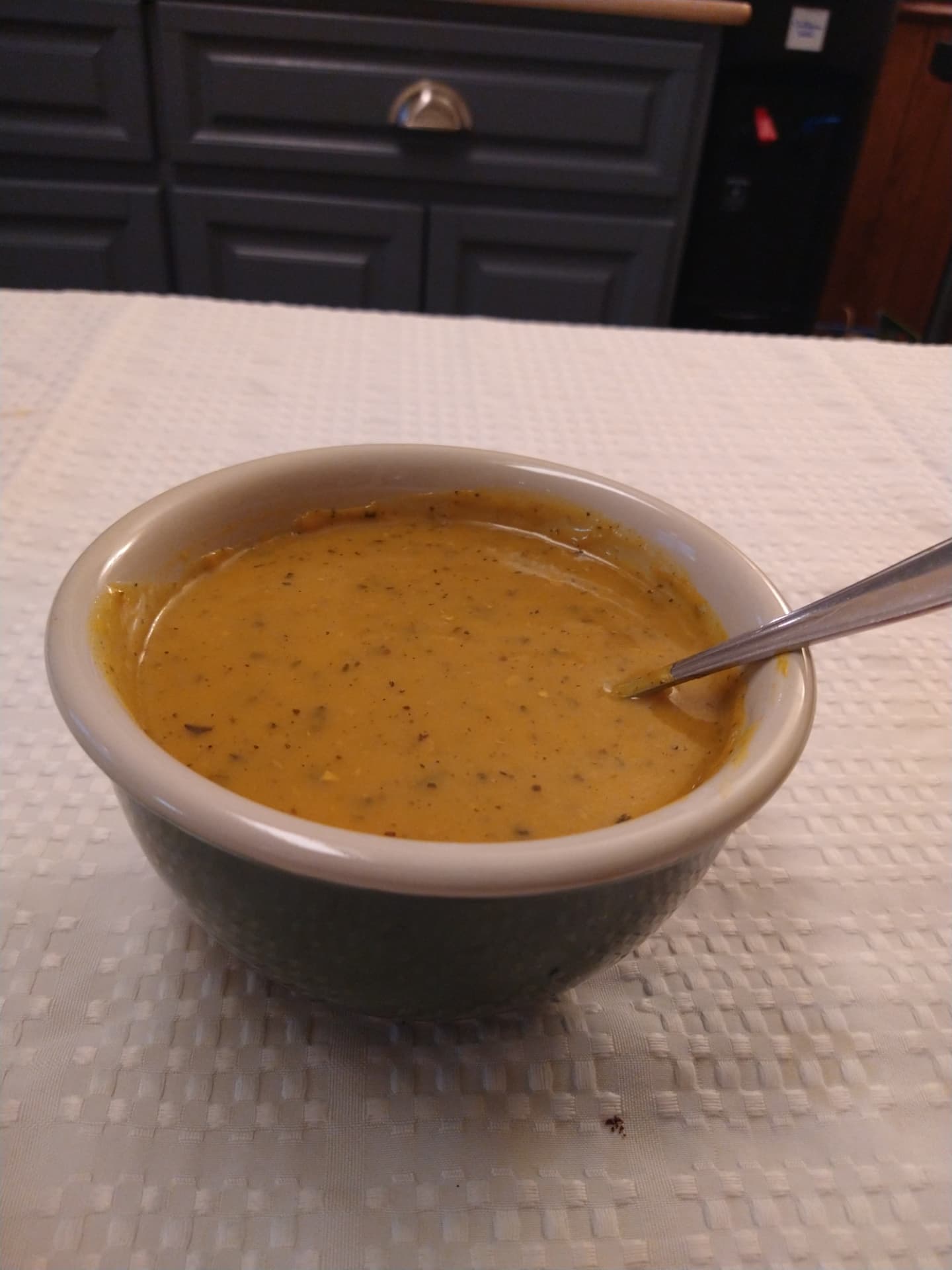 A bowl of glossy homemade gravy with flecks of seasoning and a metal spoon resting inside, set on a cream tablecloth