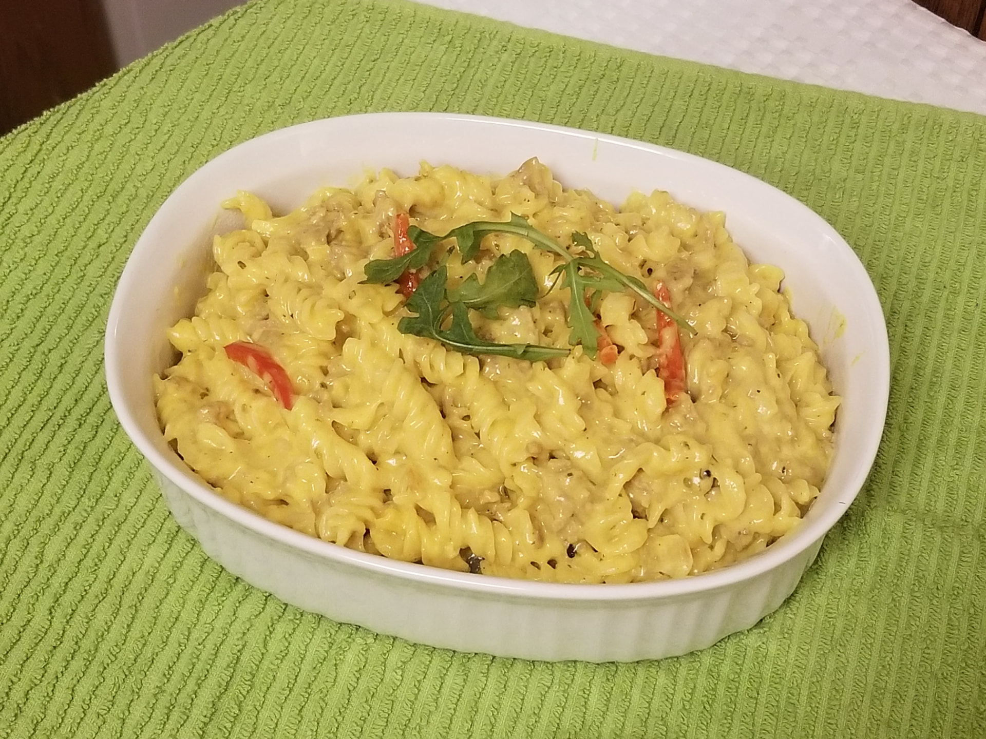 Spiral pasta baked mac and cheese in a creamy golden cheese sauce, served in a white oval baking dish and topped with red pepper strips and fresh greens