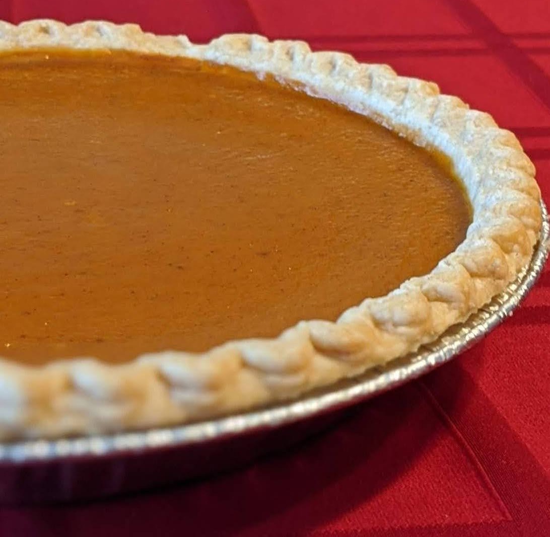 A whole sweet potato pie in a foil pie tin on a red tablecloth, with a crimped golden pastry crust